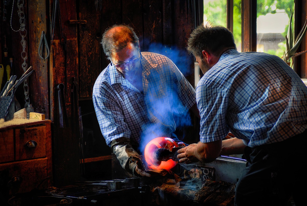 Interior of a glassblowing studio showing a glowing furnace, workbench, and rows of finished glass pieces